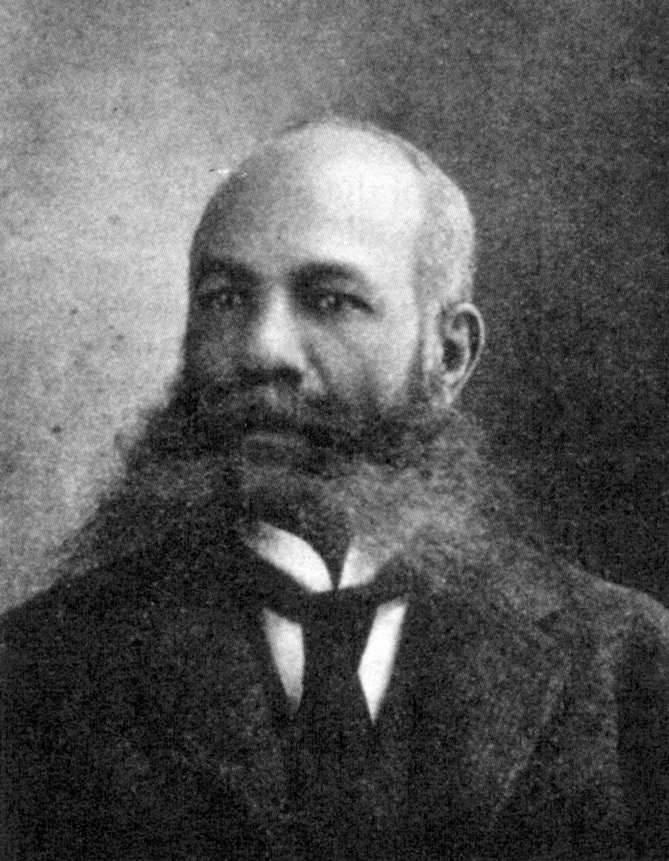 alexander miles looks to the left, he wears a suit jacket, collared shirt and tie with a large full beard