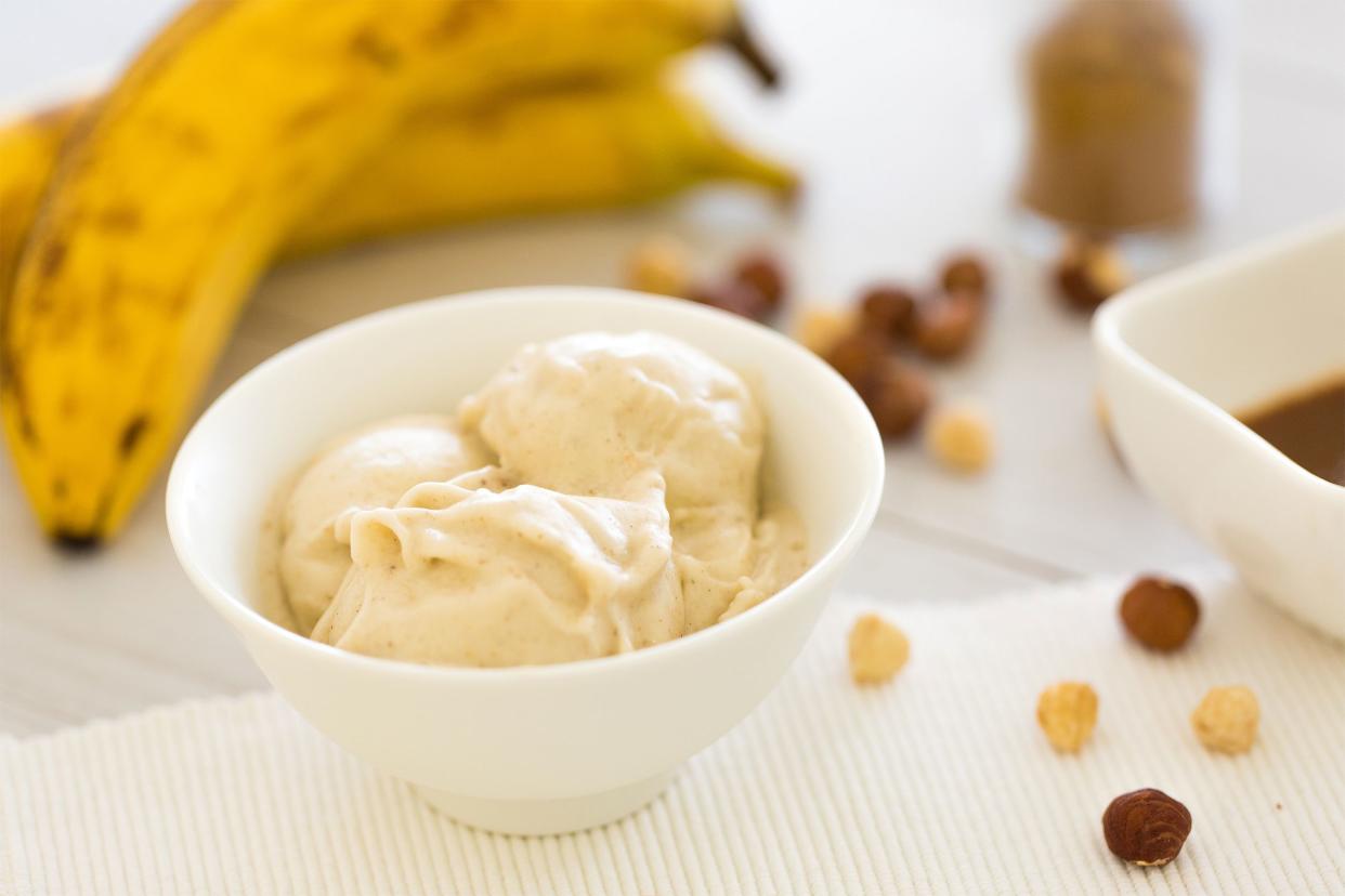 Banana ice cream in a small white bowl on a white napkin surrounded by pieces of caramel with bananas and other ingredients in the background