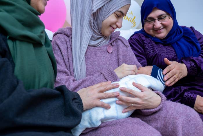 Three women, dressed in modest outfits, surround a swaddled baby as one woman tenderly holds the infant
