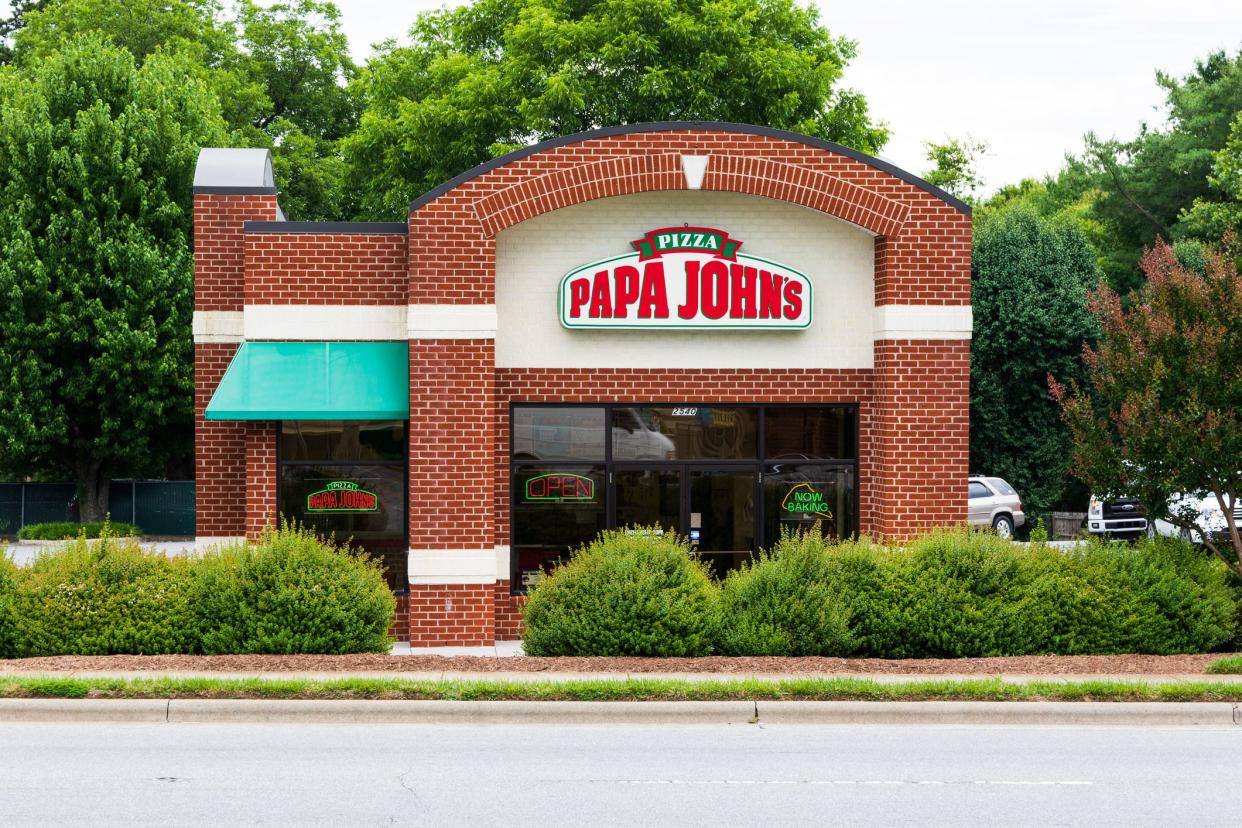 Front exterior of Papa John's pizza restaurant, Hickory, North Carolina, surrounded by a parking lot, front is lined with bushes, lots of trees in the background and a road in the foreground