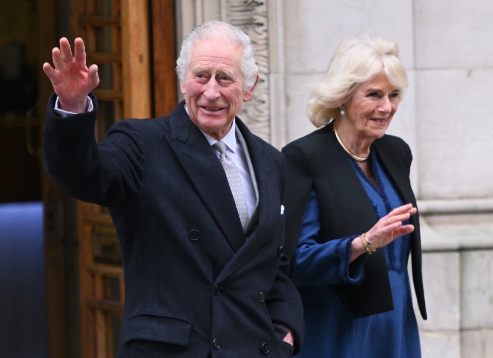 king charles iii and queen camilla wave and smile after walking out a building, he wears a dark peacoat with a purple collared shirt and silver patterned tie, she wears a black coat over a blue long sleeve dress and golden necklace