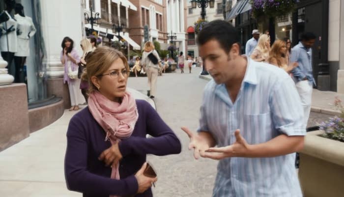 Jennifer Aniston and Adam Sandler in a casual street scene. Aniston wears a scarf and glasses and looks concerned while Sandler gestures animatedly