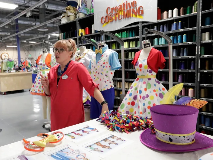 A costume designer stands next to bright costumes and a Mad Hatter character hat in a Creative Costuming studio filled with costumes and threads.