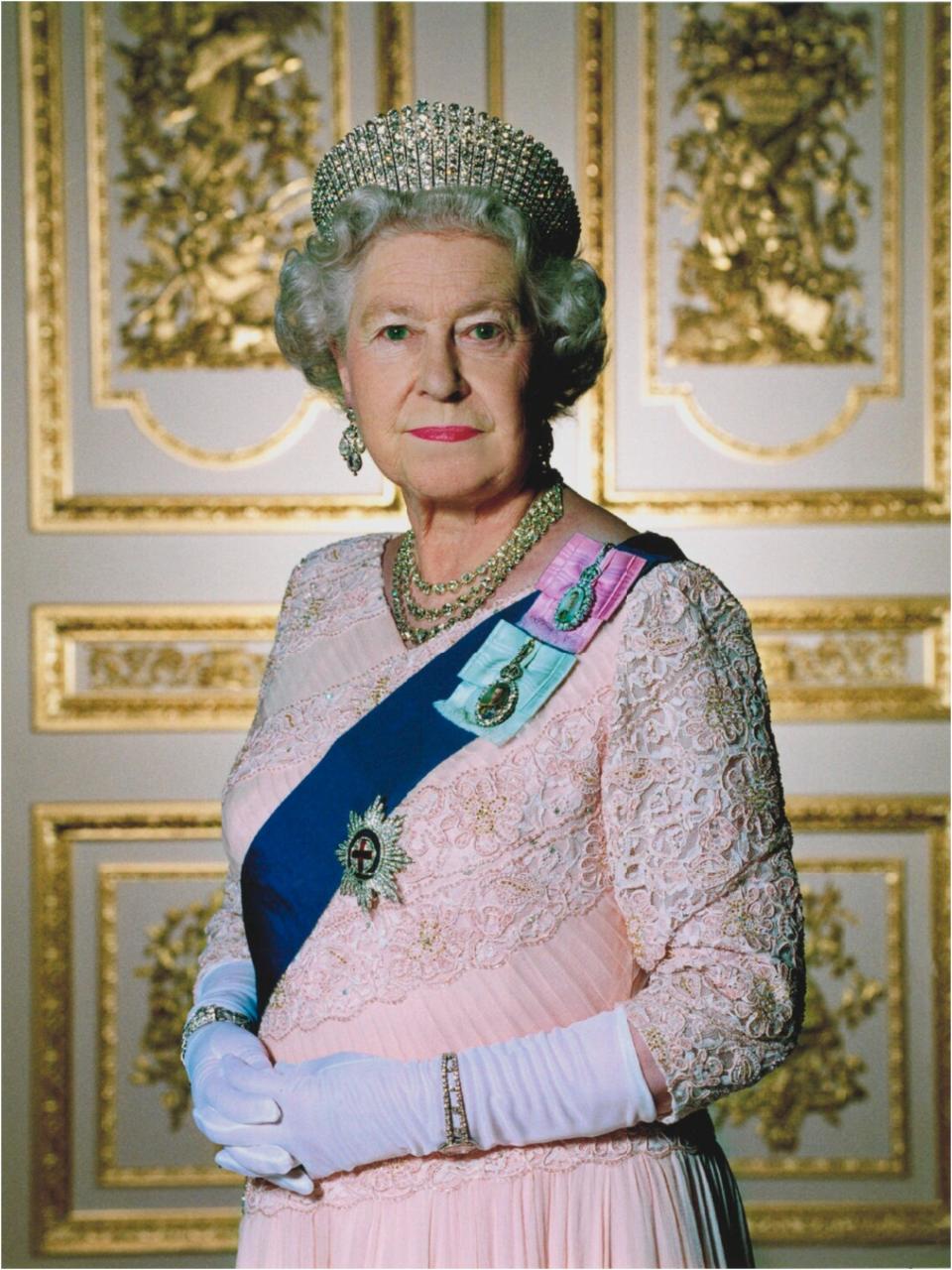 The Queen posing in a light pink lace and chiffon dress with a blue sash and medals along with gold jewelry, a large crown, and white gloves.