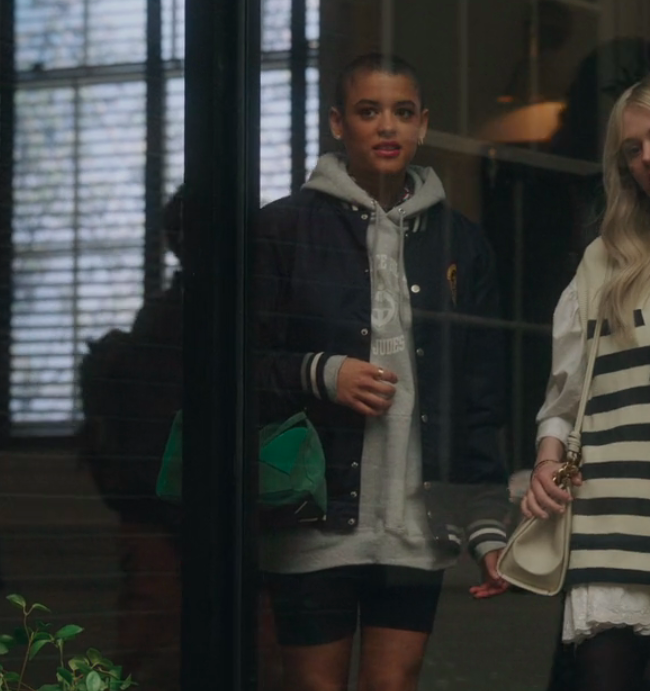 Whitney Peak and Emily Alyn Lind, dressed casually, are seen through a glass window walking indoors. Whitney wears a hoodie and jacket; Emily in a striped shirt