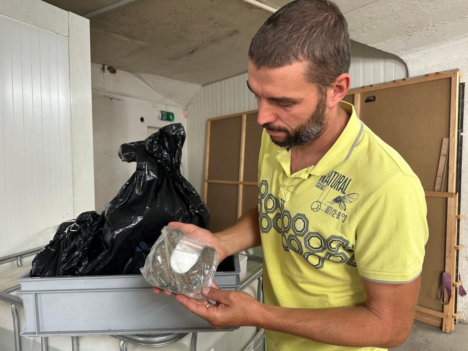 archaeologist in yellow shirt holds ancient rusty horseshoe in plastic bag beside a cart with a grey bin holding a large black trashbag in a large basement storage room