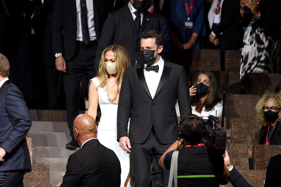 Ben Affleck, wearing a classic black tuxedo, and Jennifer Lopez, wearing a floor-length white mermaid gown, wearing masks as they enter a theater.