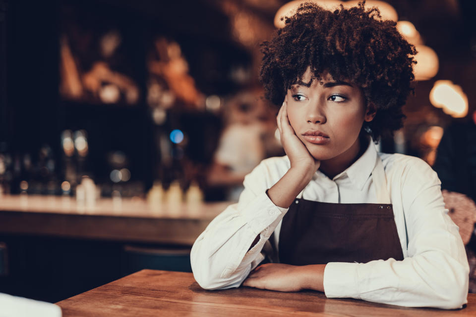 A person with curly hair, wearing a white shirt and an apron, leans on a wooden table in a restaurant, appearing deep in thought