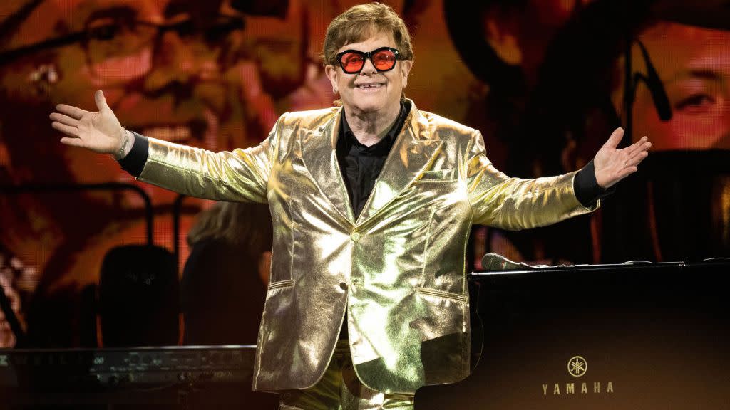 elton john smiles with his arms outstretched at his side, he wears a gold suit with a black collared shirt and pink tinted glasses with black frames