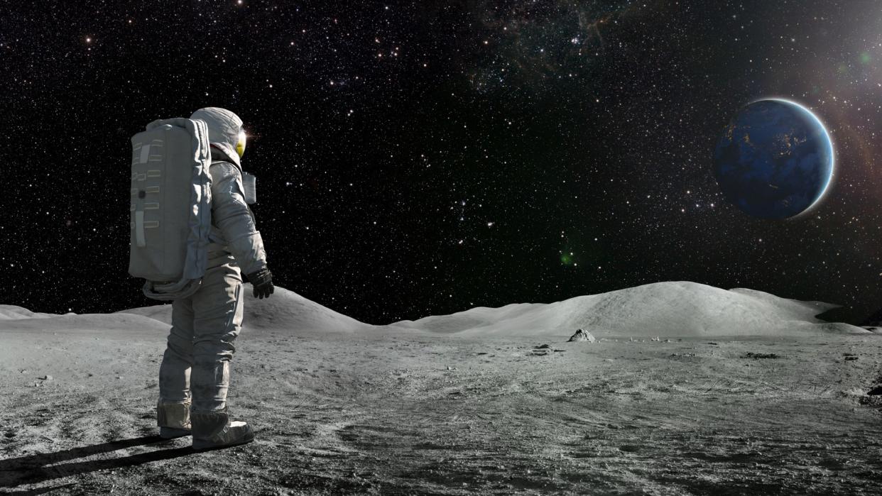  A lone astronaut standing facing away from the camera dressed in full space suit with backpack, stands still looking towards a distant planet Earth. 