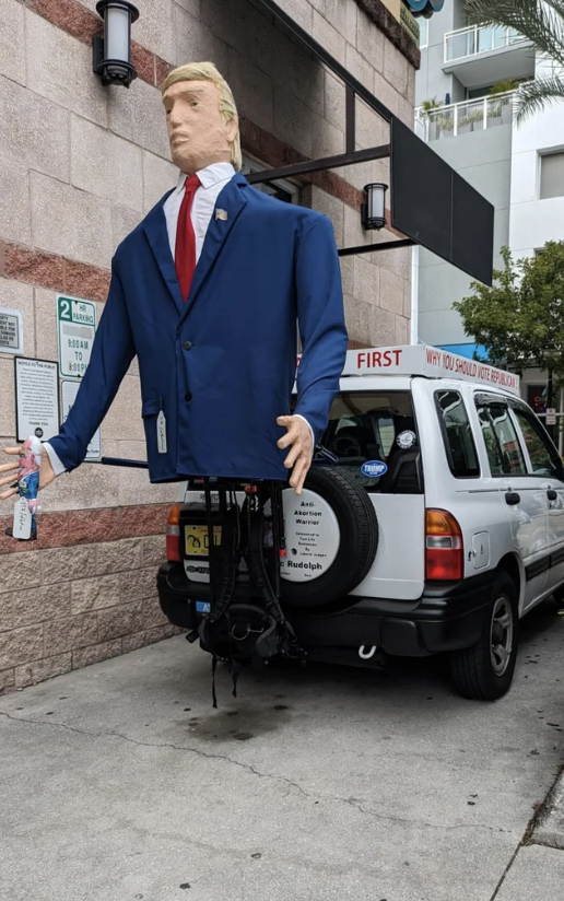 A large Donald Trump figure on a unicycle, in front of a vehicle with text and a mannequin