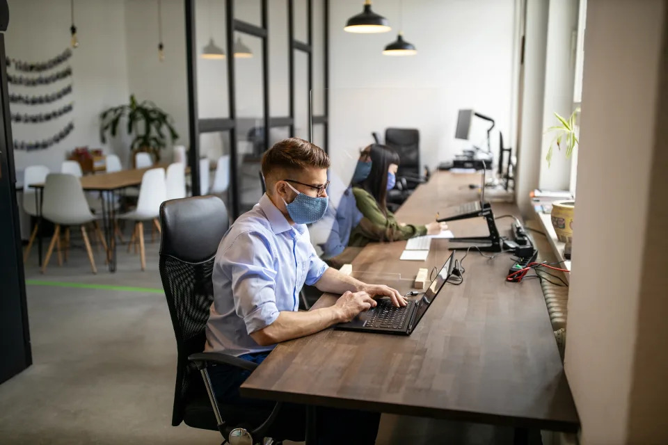 Businessman with protective face mask working at his desk. Business people return back to work after pandemic lockdown sitting at desk with protection guard between them.