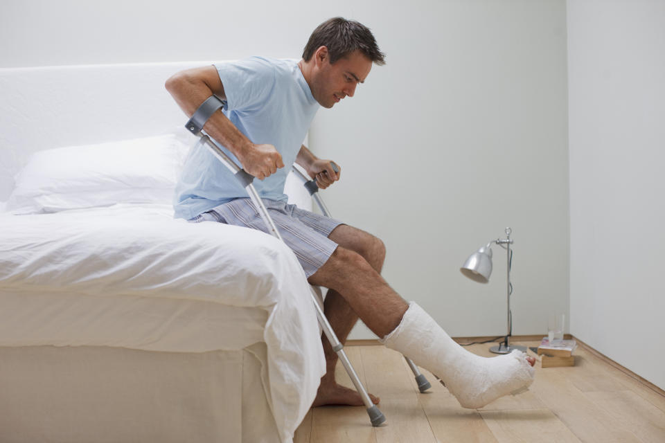 Man sitting on edge of bed with leg in cast, trying to hoist himself up with crutches