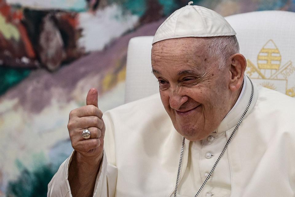 Pope Francis, squinting slightly, gives a thumbs-up and smiles. He is seated on a white chair with what appears to be a papal seal in front of a colorful backdrop.