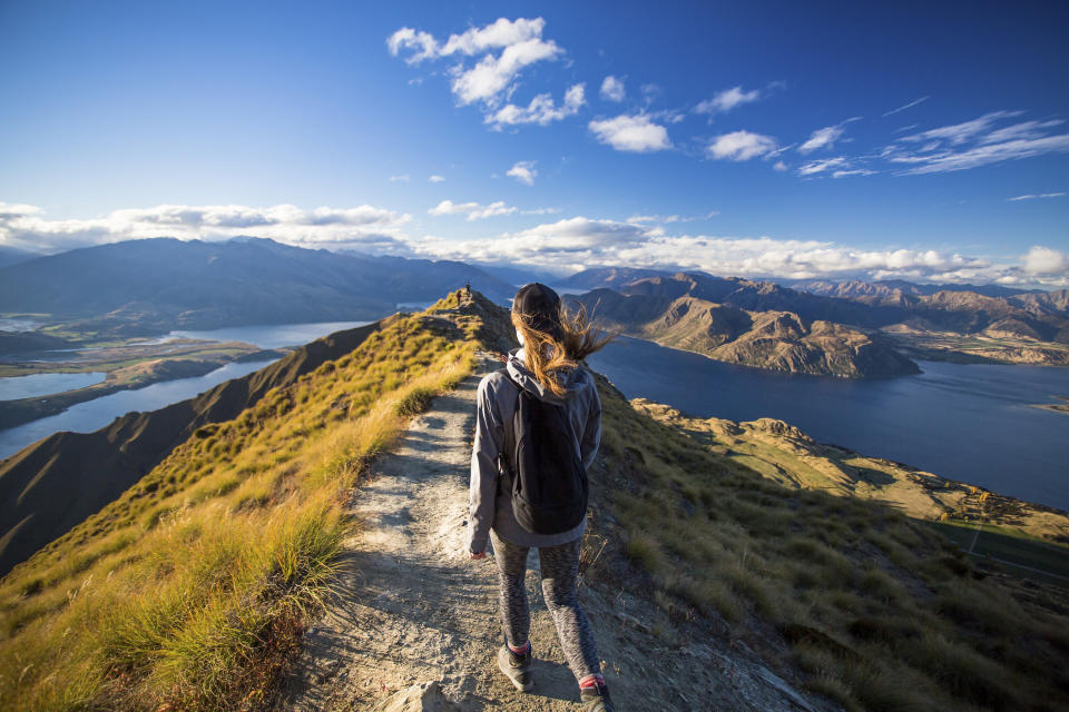 A person with a backpack walks along a mountain trail overlooking a lake and surrounding hills, representing adventure and outdoor exploration