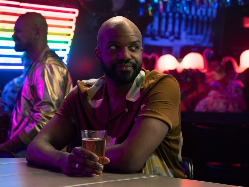 carl clemons-hopkins as marcus in hacks, wearing a brown shirt, and sitting at a bar holding a beer. behind them, there's a neon pride flag light and lit-up plastic flamingos