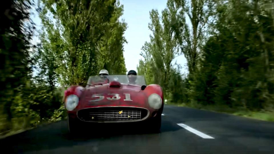 a vintage ferrari approaches the camera along a narrow tree lined road in a still from michael mann's upcoming film