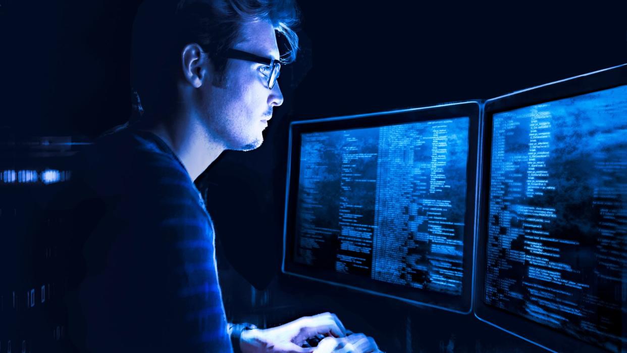  young man wearing glasses and standing in front of a panel of large computer monitors displaying code 