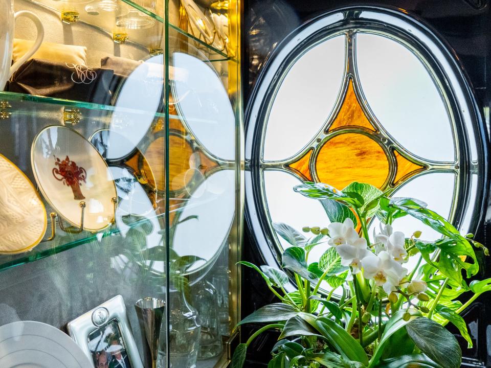 A glass box displaying dishes on the left and a stained-glass window with an orange triangle in the middle behind a bouquet of white flowers and green leaves on the right