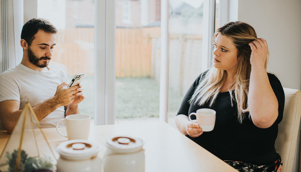 Two people seated at a table with one looking at a phone and the other looking at them while holding a mug