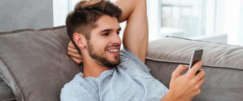 Photo of happy young man lies on sofa and looking on phone