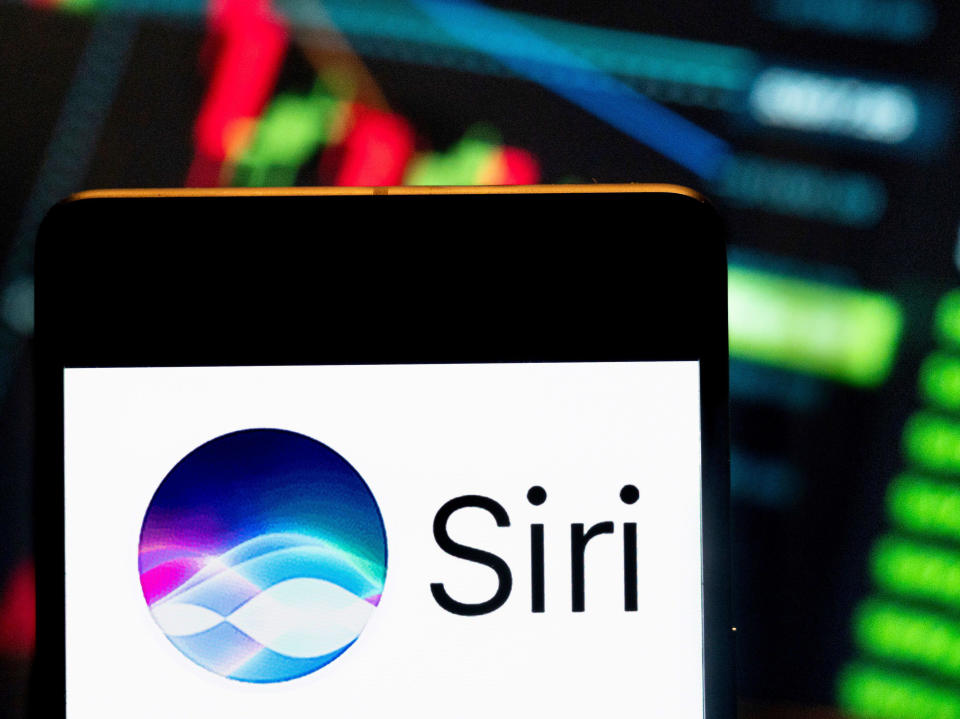 In this photo illustration, the digital assistant owned by Apple, Siri, logo seen displayed on a smartphone with an economic stock exchange index graph in the background.