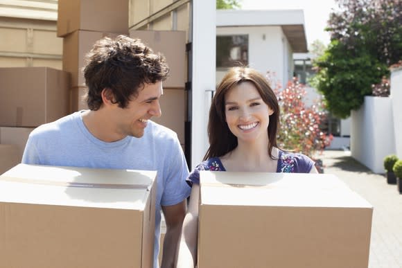 A young woman and man carrying boxes with a truck full of boxes and a house behind them.