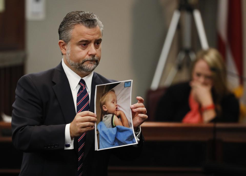 Defense attorney Maddox Kilgore holds a photo of Cooper Harris in this 2016 file photo during a murder trial for Cooper's father Justin Ross Harris, who was accused of intentionally killing him in June 2014 by leaving the child in the car. Cooper’s mother Leanna Taylor weeps on the witness stand in the background of the Brunswick, Georgia, courtroom. [AP Photo/John Bazemore}