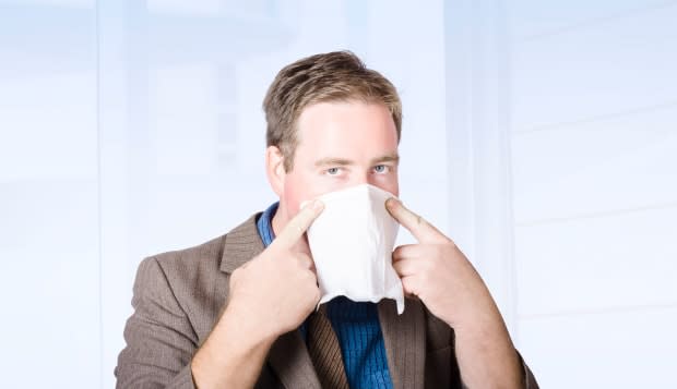 Unwell male office worker suffering from a contagious virus, covering face with tissue during cold and flu season