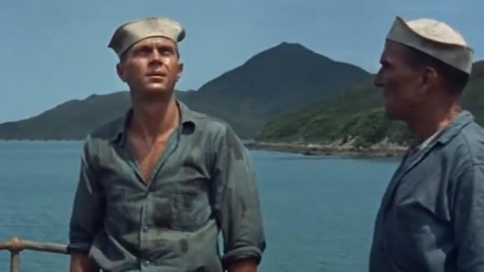 Steve McQueen in a Navy uniform standing on the bow of a ship with the ocean and an island behind him