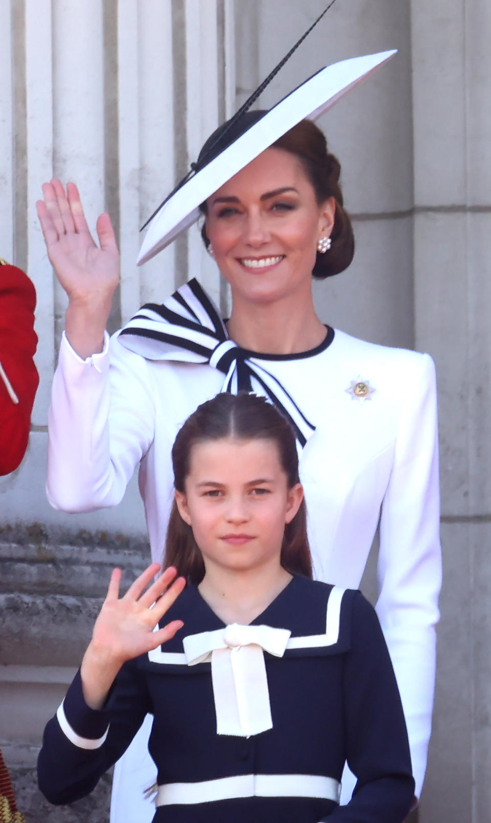 Kate Middleton in formal attire and fascinator hat, accompanied by Princess Charlotte in a navy dress with a white bow, both waving