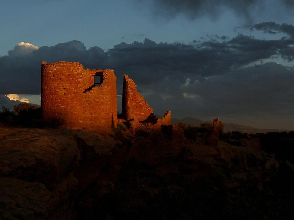 Hovenweep castle ruins are shown bathed in bright orange sunset light set against a stormy sky