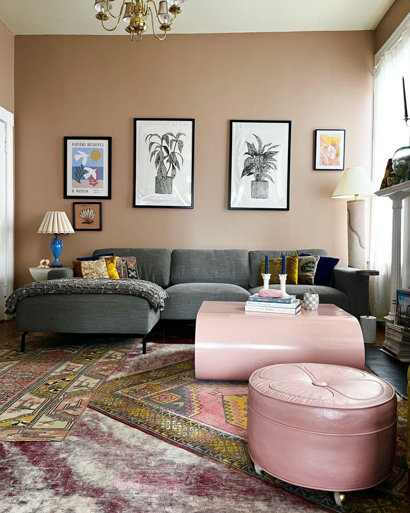 Gray couch and pink coffee table and ottoman sit atop multiple area rugs in living room with plant artwork hung on pink brown wall.