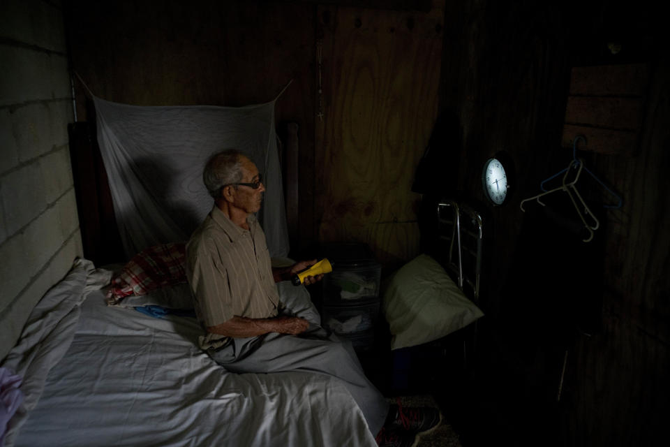 En esta fotografía del 8 de septiembre de 2018, Ramón Alicea Burgos utiliza una linterna para leer la hora en su reloj de pared en una habitación temporal de madera que construyó bajo la estructura de su casa parcialmente reconstruida, sin terminar y sin electricidad por falta de dinero, en el pueblo montañoso de Barranquitas, Puerto Rico. Burgos, de 82 años, dijo que un médico local está recolectando donaciones de otros médicos y pacientes para ayudarlo a recaudar fondos para terminar su casa. (AP Foto/Ramón Espinosa)