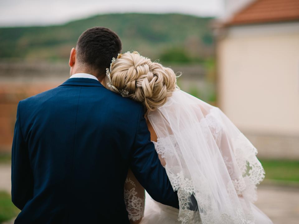 Groom gently hugs the bride from the back on a beautiful background.