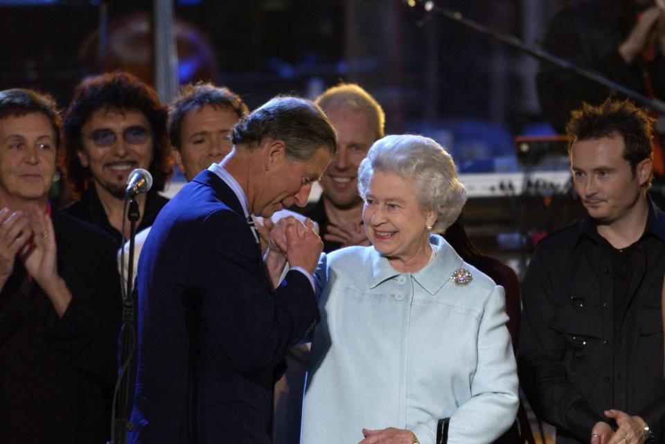 prince charles, wearing a blue suit, kissing the hand of queen elizabeth ii, wearing a blue dress, standing on a stage with several men watching and clapping