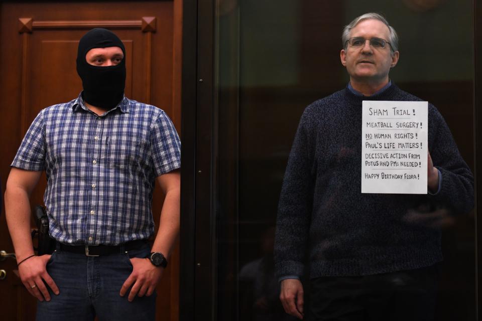 Paul Whelan stands behind glass holding a sign that reads: Sham trial! Meatball surgery! No human rights! Paul's life matters! Decisive action from POTUS and PMs needed! Happy Birthday Flora!