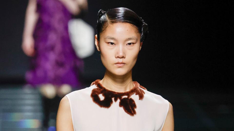 A model walks the runway during the Prada Ready to Wear show wearing a white dress with brown floral accents.