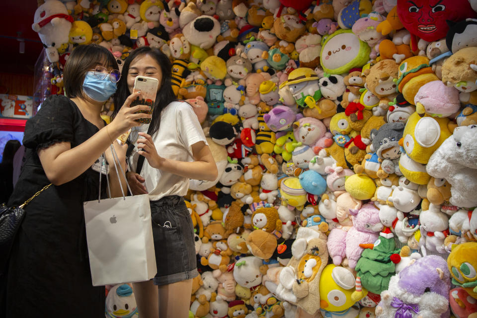 A woman wearing a face mask to protect against the coronavirus poses for a selfie in front of a wall of stuffed animals at an arcade in Beijing, Saturday, Aug. 1, 2020. China reported a more than 50% drop in new cases of COVID-19 on Saturday in a possible sign that its latest major outbreak in the northwestern region of Xinjiang may be waning. (AP Photo/Mark Schiefelbein)