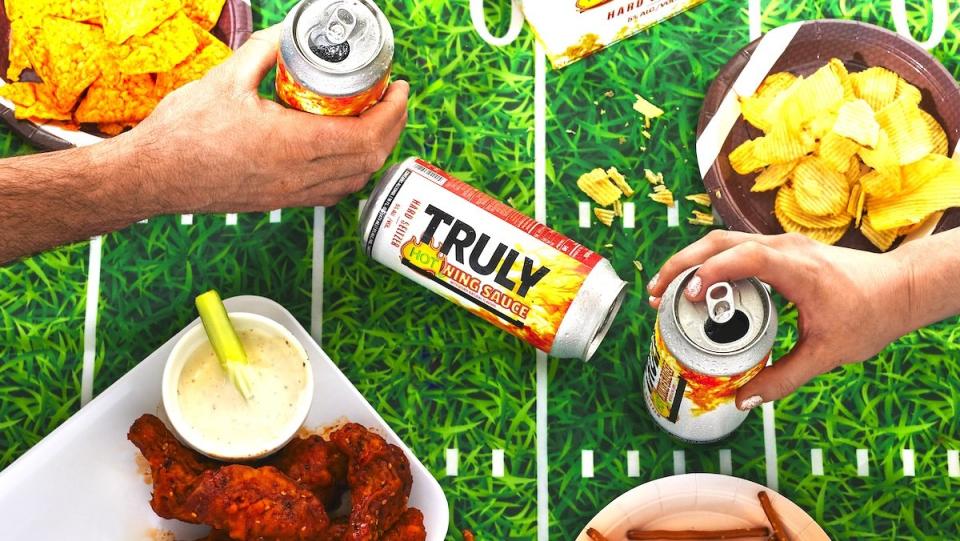 Cans of Truly Hot Wing Sauce Seltzer on a table with human hands and buffalo wings near by