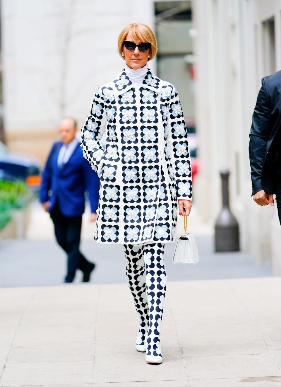 Celine in a knee length coat and matching tights. The white ensemble has an all-over black and light blue square flower pattern. She's wearing black round sunglasses and white ballet flats.