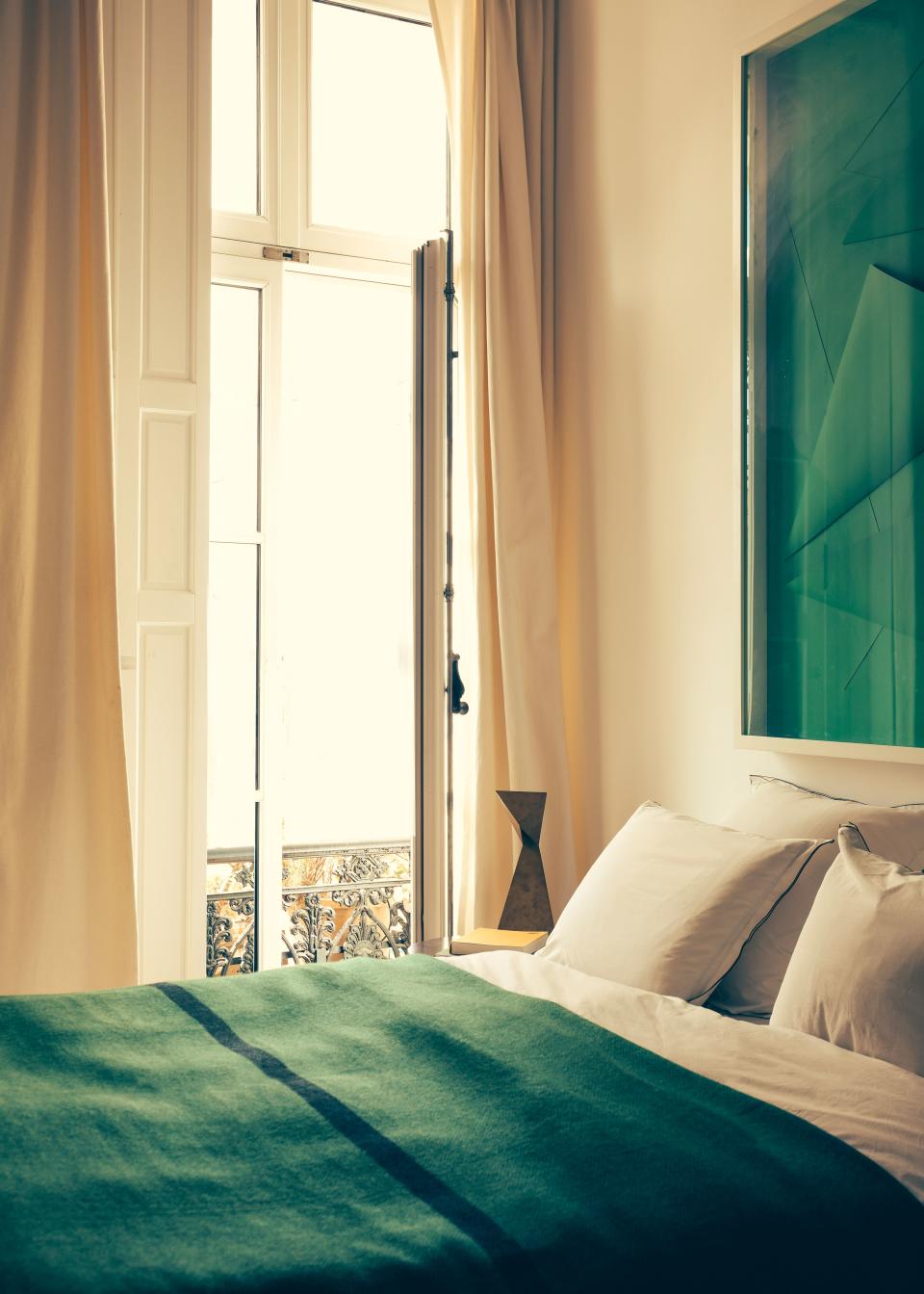 The master bedroom looks out over gardens, and the cool interior is punctuated with pops of color. The green bedcover is handwoven in Tibet, and the painting is by British/French artist Marine Hugonnier. The bedside lamp is by Champsaur, called Origami by Pouenat Ferronnier.