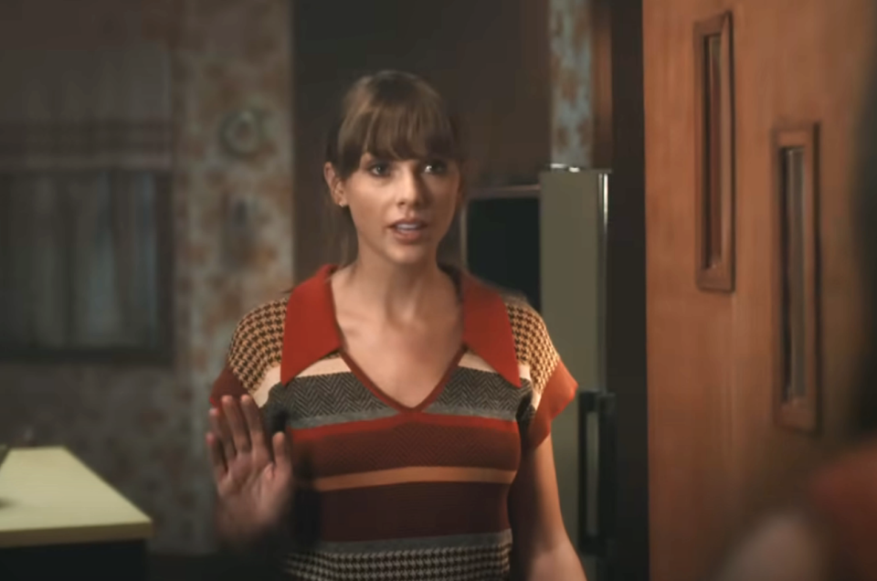 Woman in striped dress stands in a kitchen, looking surprised with hands up