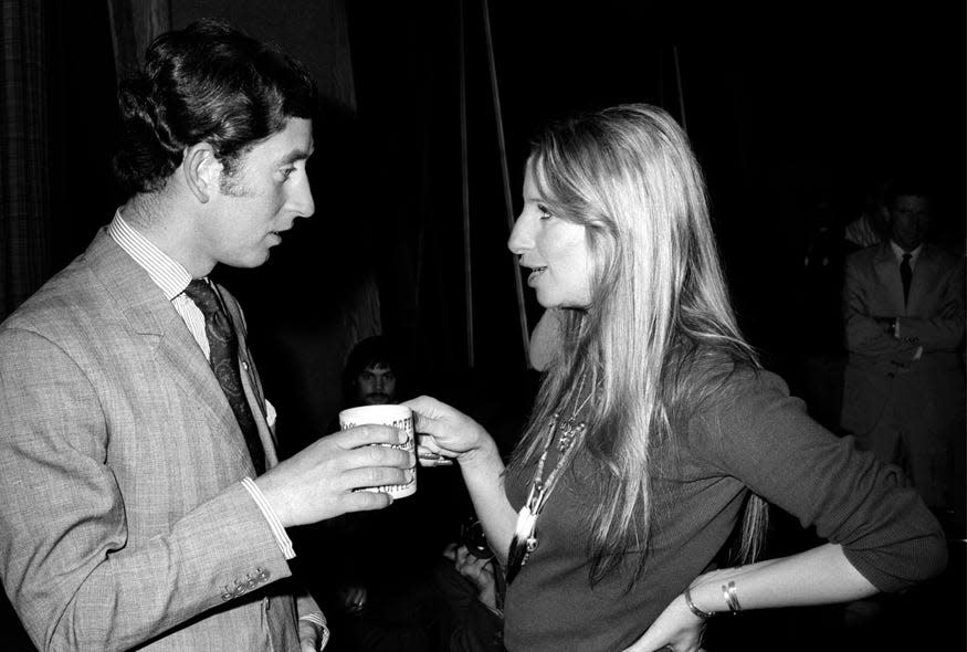 A young King Charles III, wearing a suit and tie, stands talking to a young Barbra Streisand, wearing a long-sleeved shirt and holding a coffee mug, in this black and white photo from 1974.