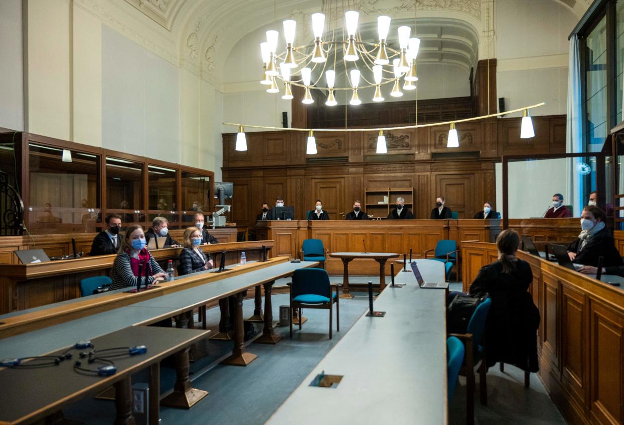 Participants in a trial, sitting at a long tables and wearing surgical masks, appear before a panel of judges in a wood-paneled courtroom..