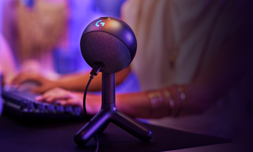 Marketing image of the Logitech G Yeti Orb mic. It sits in the foreground on a desk as a gamer in the background (blurred) rests their hands on a keyboard.