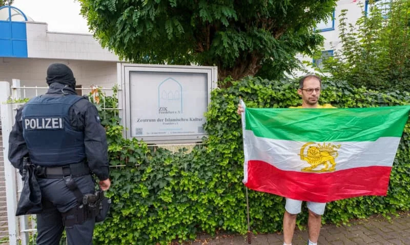 A man stands with an old Persian national flag next to a police officer and a display case. Following the ban on the nationwide Islamic Center Hamburg (IZH), three properties in Frankfurt am Main and Bad Homburg have been searched by the police. Andreas Arnold/dpa