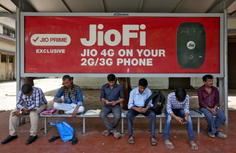 FILE PHOTO: Commuters use their mobile phones as they wait at a bus stop with an advertisement of Reliance Industries' Jio telecoms unit, in Mumbai