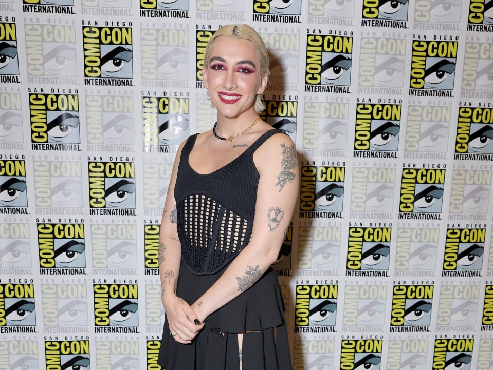 mason alexander park on the comic con red carpet, wearing a black dress with a laced midriff section and skirt made out of loosely stiched together black rectangles. they have blonde hair and prominent pink makeup