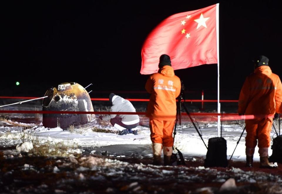 A picture shows the burnt up capsule from China's Chang'e 5 mission laying in a snowy field at night. In the forefront are a Chinese flag and two workers dressed warming in orange work garb. Someone is inspecting the probe which is within a cordoned off area.